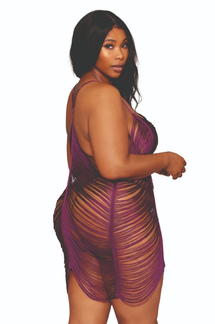 A woman with long, dark hair is wearing the Flirty Plus Size Draped Fringe Chemise in a vibrant purple hue. She poses confidently against a plain white background, showcasing the wavy, fringed design and thin straps. The fence-net design of the chemise highlights her style as she stands poised and elegant.