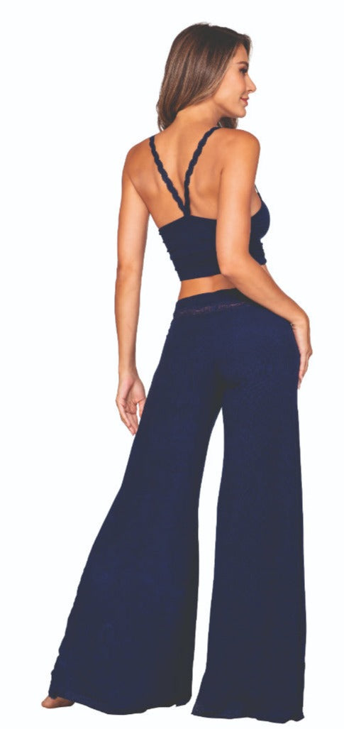 A woman is modeling the Comfortable Rib Knit Jersey Sleep Pant Set in navy blue, which includes a cropped top with thin straps and high-waisted, wide-leg pants. The outfit is adorned with lace accents on both the top and pants. She stands with one hand resting on her thigh and the other by her side, smiling, ready to sleep in style.